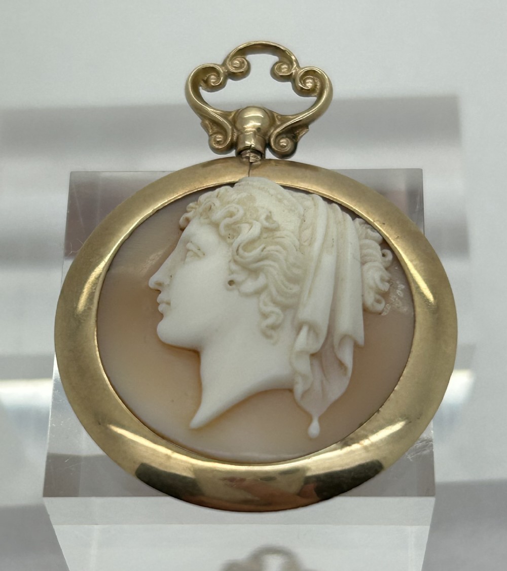 An antique round shell cameo depicting a carved female classical profile in cream set against a tan background, encased in a smooth gold frame with a top scroll bail.