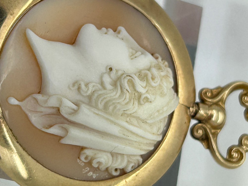 An antique round shell cameo depicting a carved female classical profile in cream set against a tan background, encased in a smooth gold frame with a top scroll bail. View V