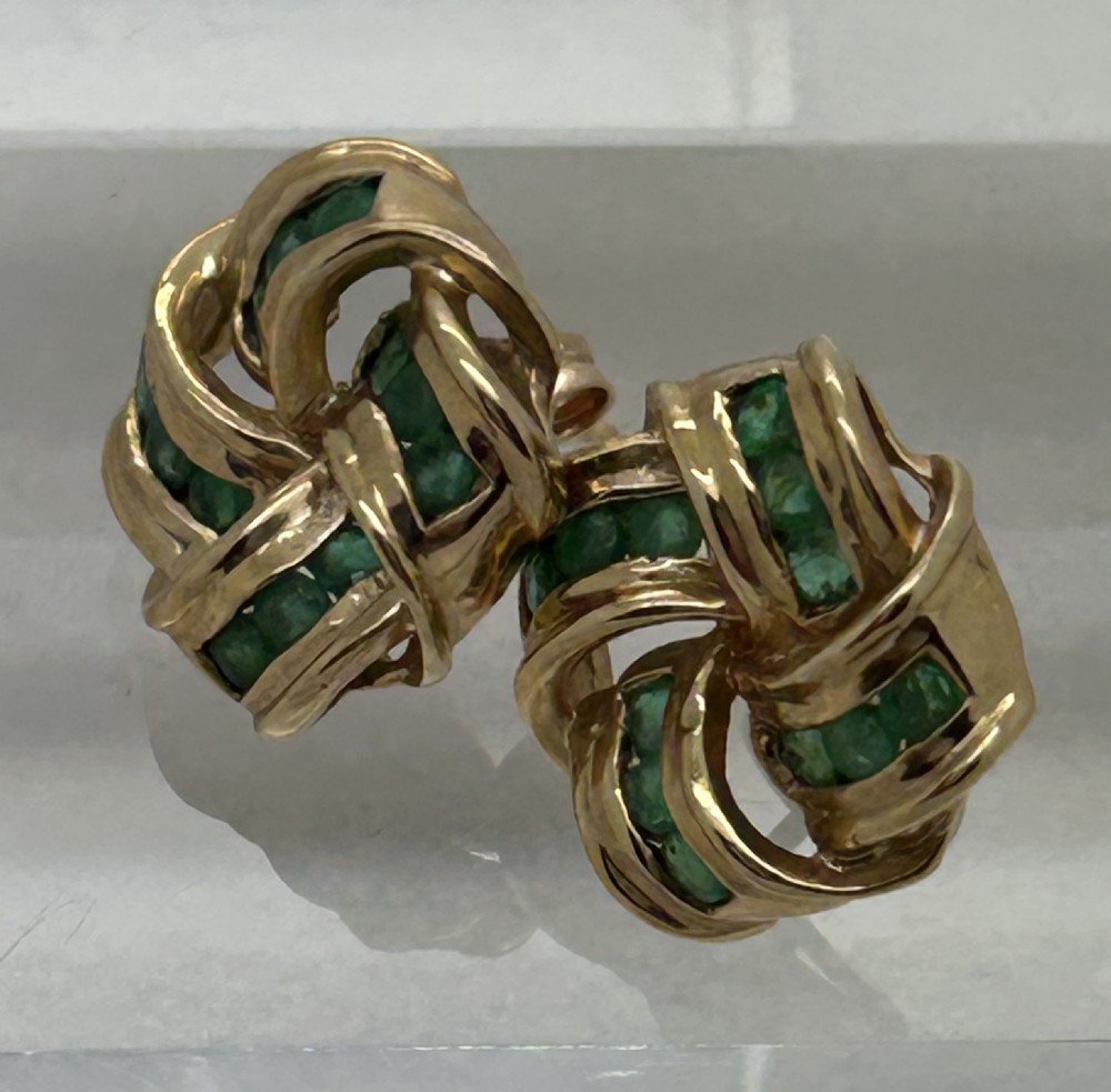 A pair of yellow gold knot earrings featuring channel-set green emerald gemstones woven into the metal bands.