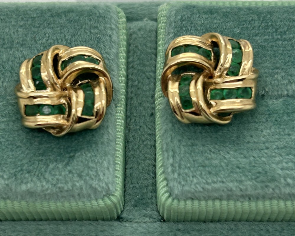 A pair of yellow gold knot earrings featuring channel-set green emerald gemstones woven into the metal bands. View II
