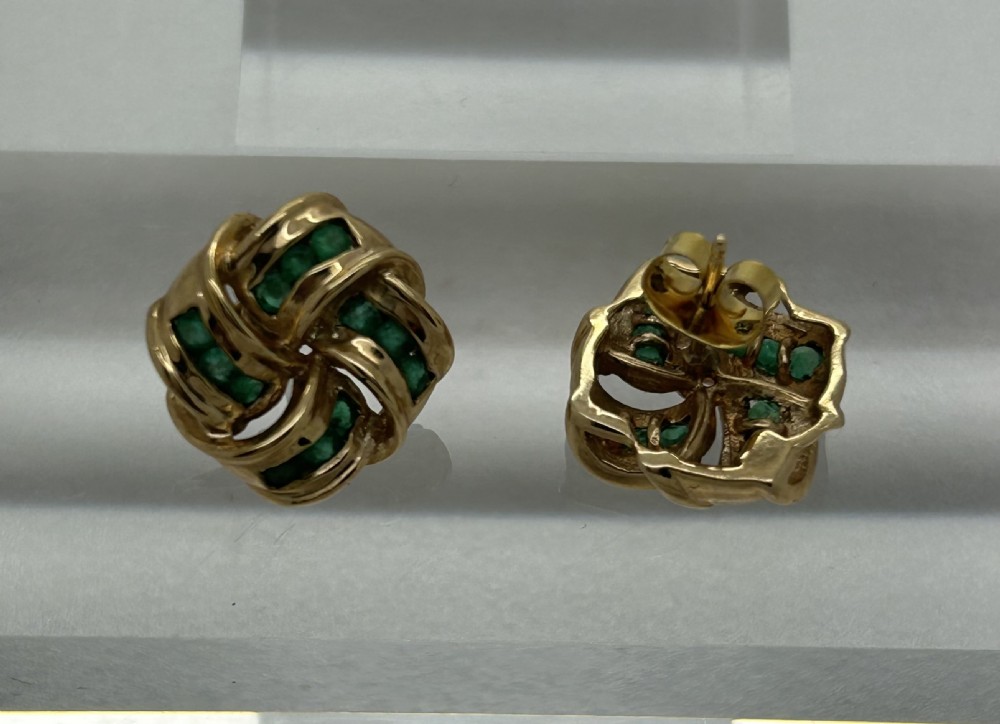 A pair of yellow gold knot earrings featuring channel-set green emerald gemstones woven into the metal bands. View V
