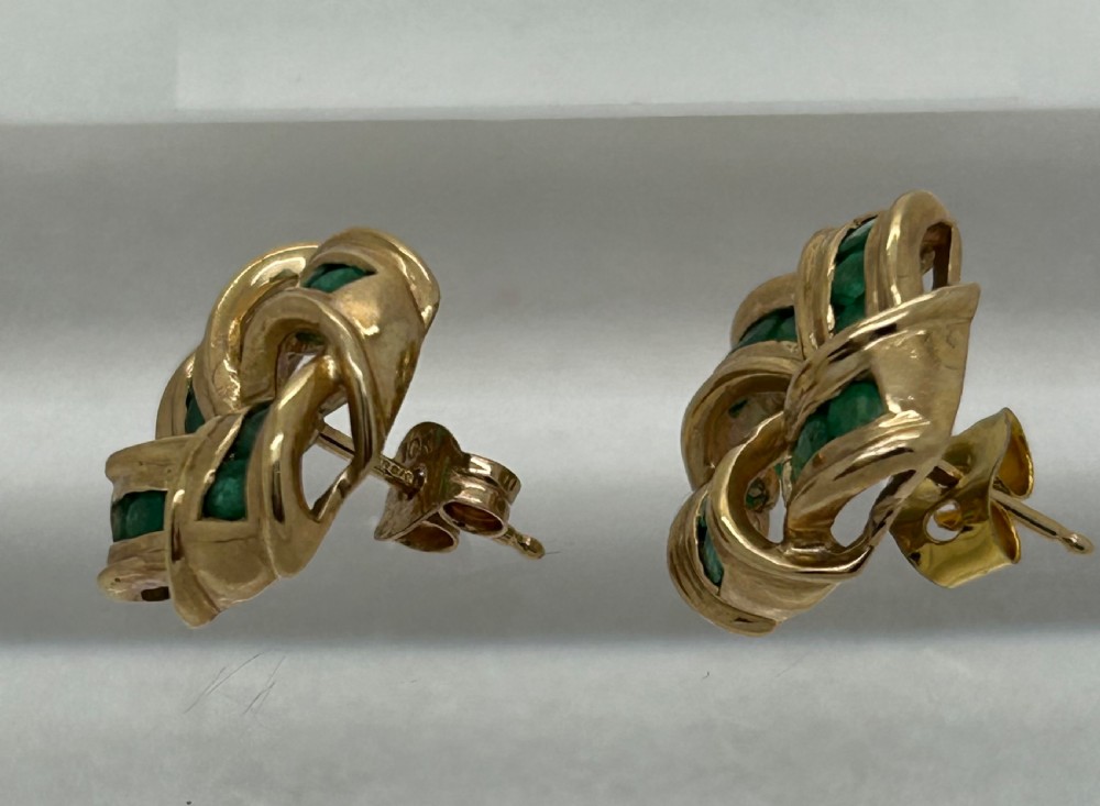A pair of yellow gold knot earrings featuring channel-set green emerald gemstones woven into the metal bands. View VI