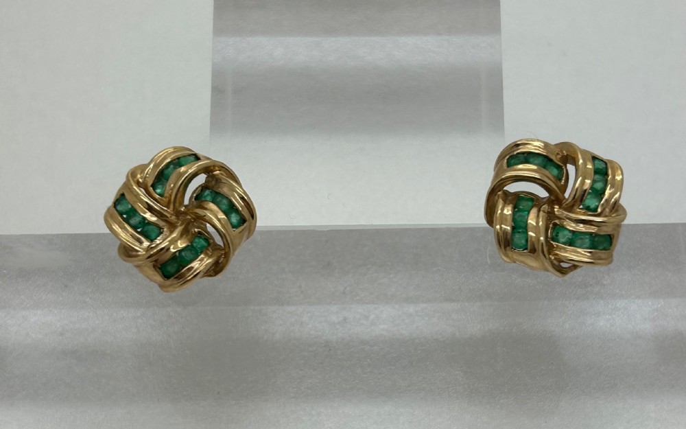 A pair of yellow gold knot earrings featuring channel-set green emerald gemstones woven into the metal bands. View VII