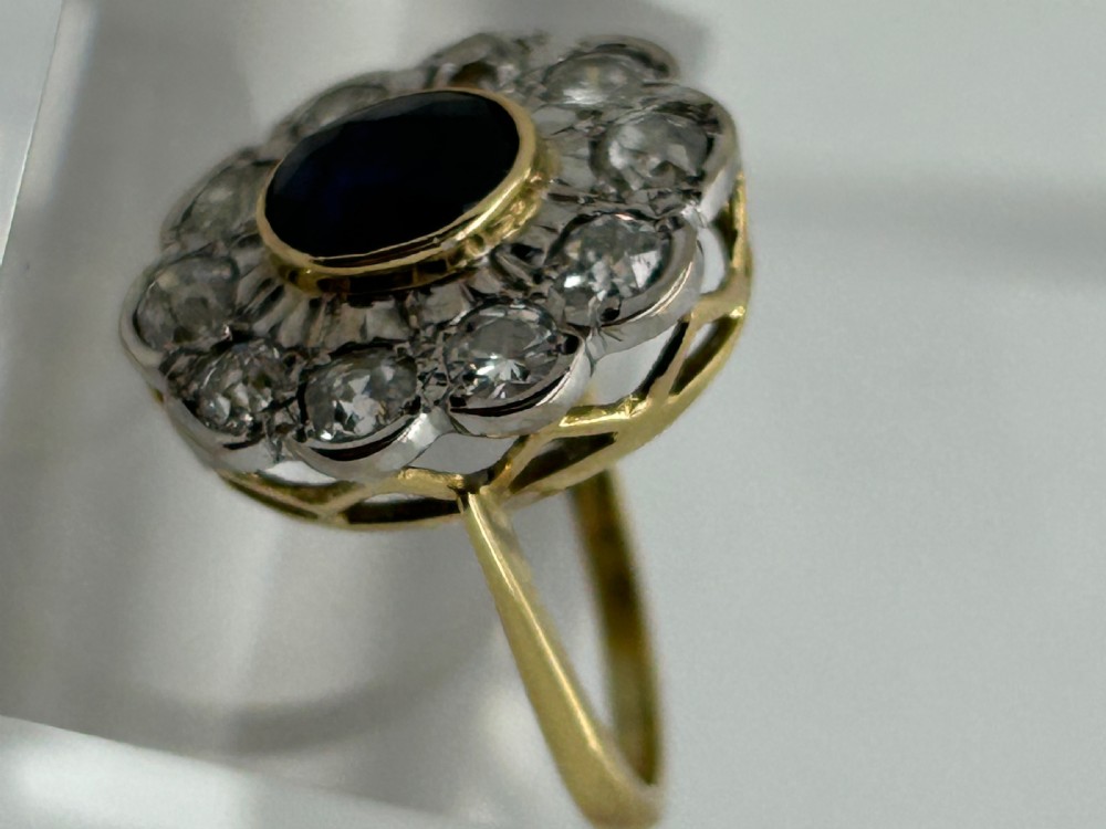 A yellow gold cluster ring featuring a central oval dark blue sapphire framed by a scalloped halo of round cut diamonds. View II