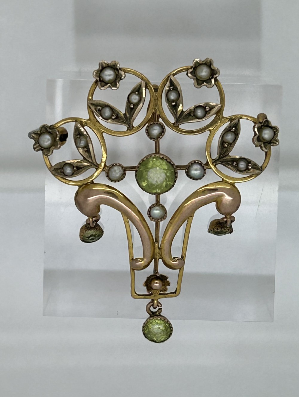 An Edwardian gold openwork pendant brooch featuring open scrolls, small seed pearls, a central faceted green peridot, and three dangling peridot droplets.