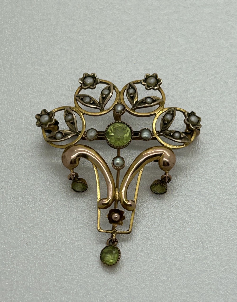 An Edwardian gold openwork pendant brooch featuring open scrolls, small seed pearls, a central faceted green peridot, and three dangling peridot droplets. View II