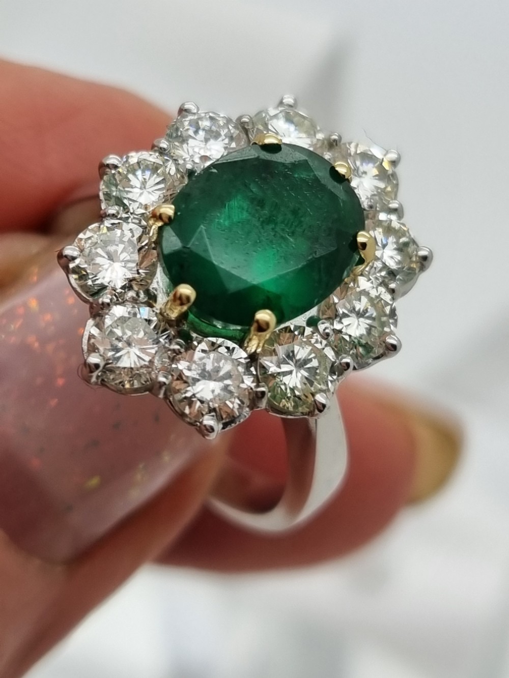 An 18ct gold ring featuring a central oval green emerald framed by a halo of round brilliant-cut diamonds. View II