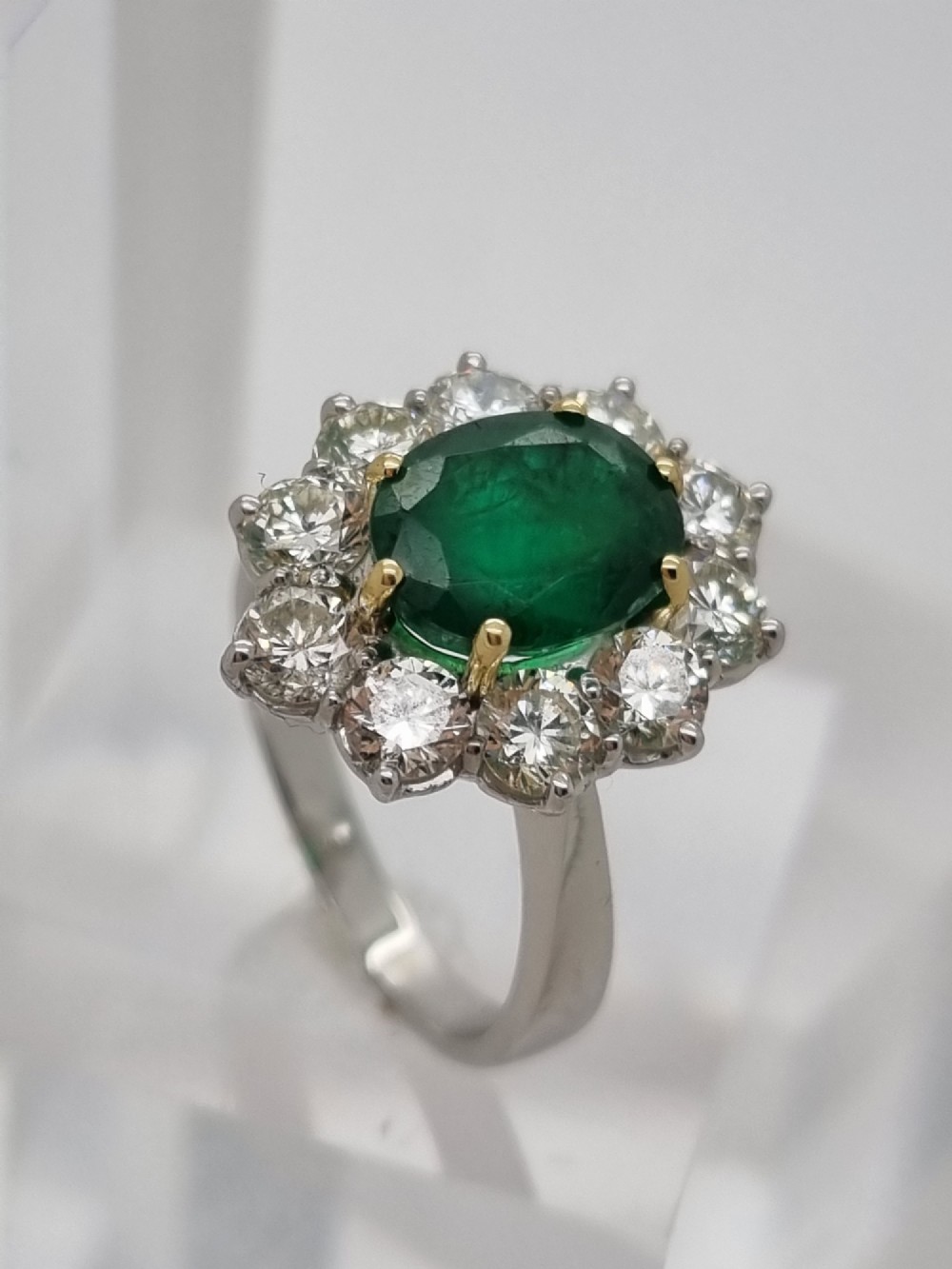 An 18ct gold ring featuring a central oval green emerald framed by a halo of round brilliant-cut diamonds. View V
