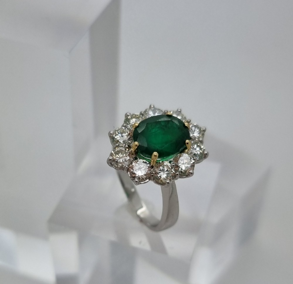 An 18ct gold ring featuring a central oval green emerald framed by a halo of round brilliant-cut diamonds. View VI