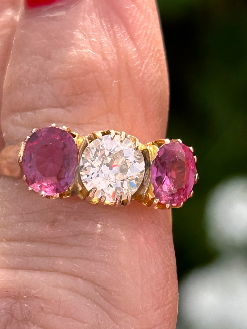 A Victorian yellow gold trilogy ring featuring a central round diamond flanked by two oval pinkish-red rubies in claw settings. View IV
