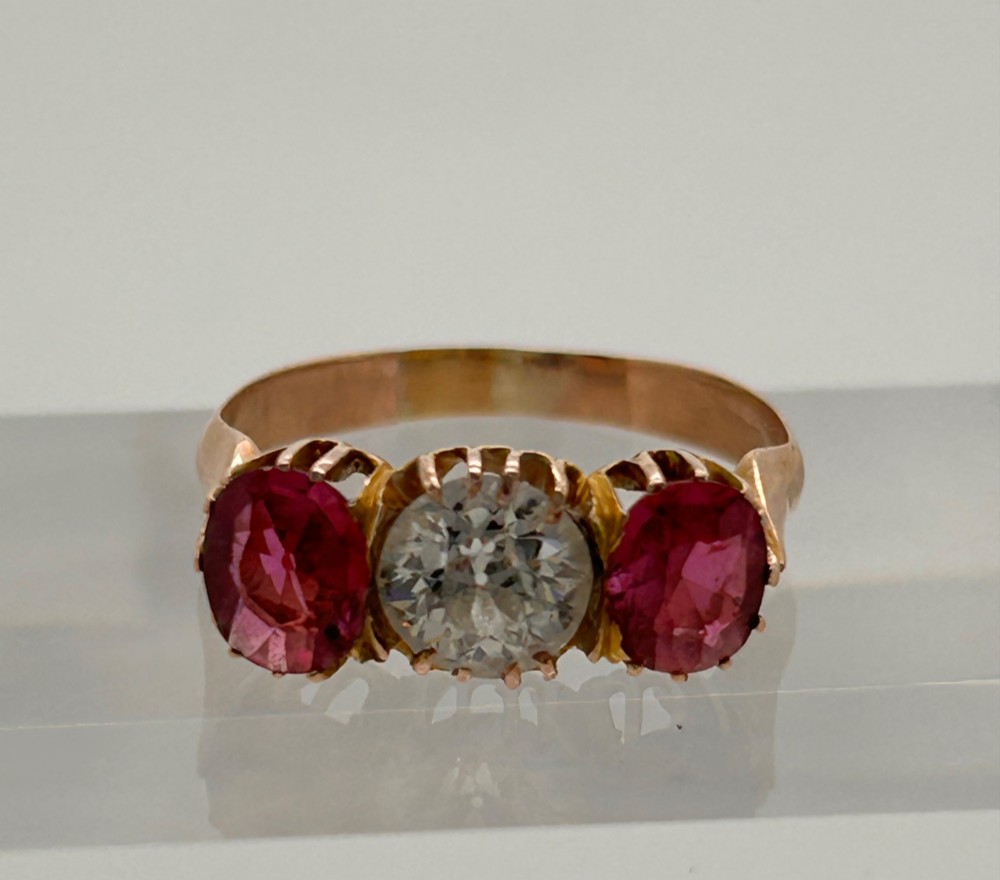A Victorian yellow gold trilogy ring featuring a central round diamond flanked by two oval pinkish-red rubies in claw settings. View V