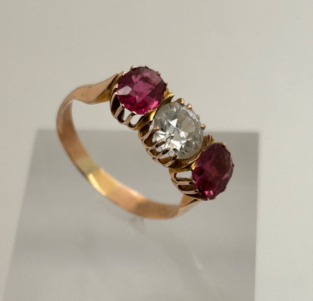 A Victorian yellow gold trilogy ring featuring a central round diamond flanked by two oval pinkish-red rubies in claw settings. View VI
