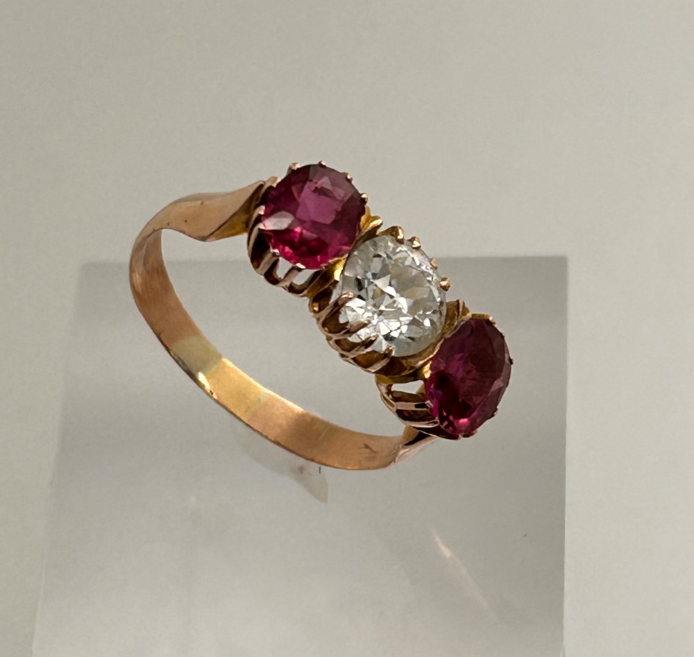 A Victorian yellow gold trilogy ring featuring a central round diamond flanked by two oval pinkish-red rubies in claw settings. View VII
