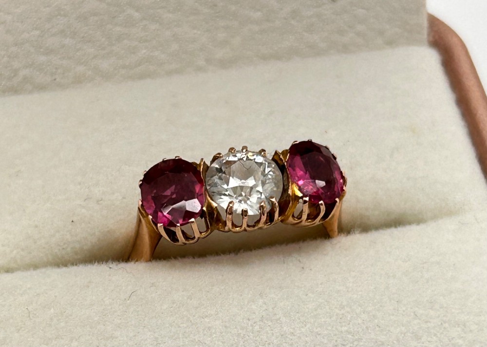 A Victorian yellow gold trilogy ring featuring a central round diamond flanked by two oval pinkish-red rubies in claw settings. View VIII