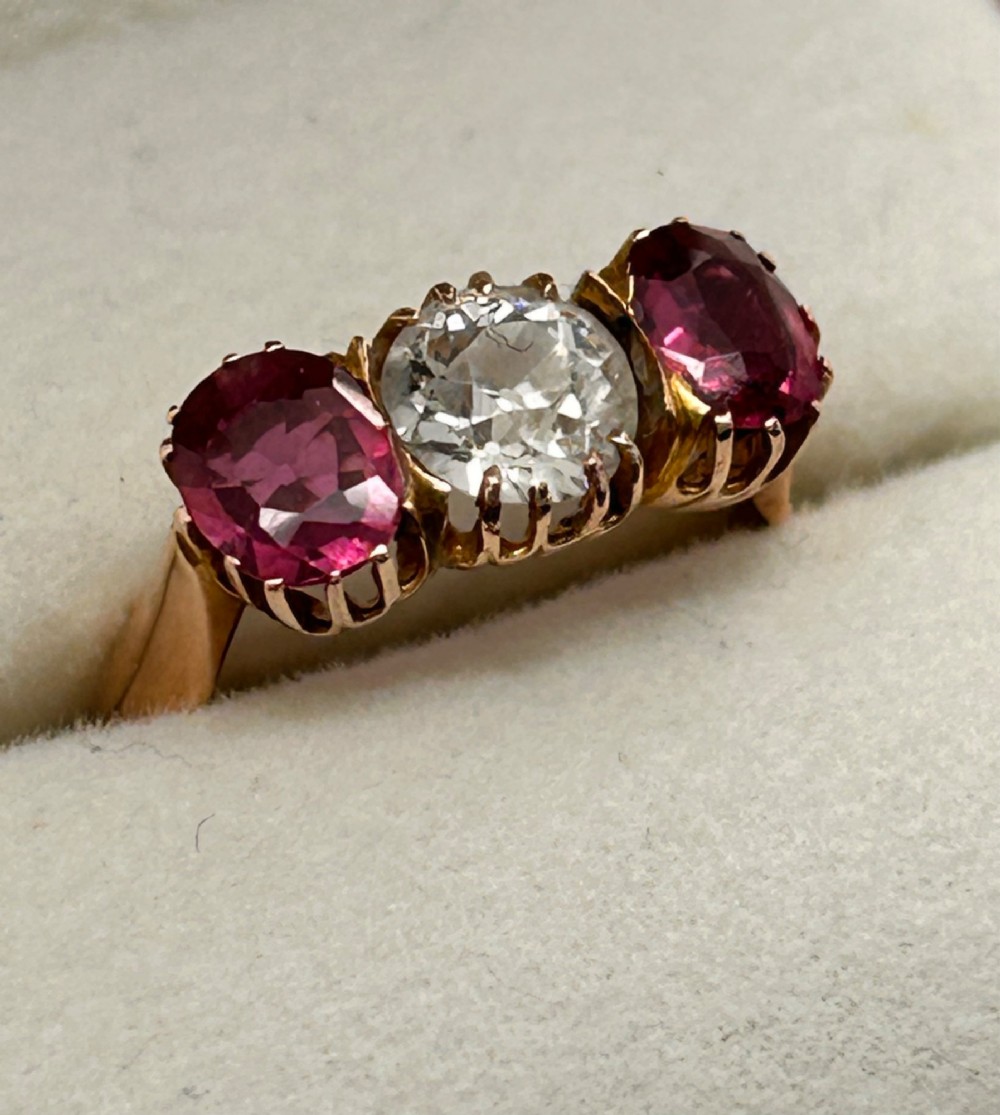 A Victorian yellow gold trilogy ring featuring a central round diamond flanked by two oval pinkish-red rubies in claw settings. View IX