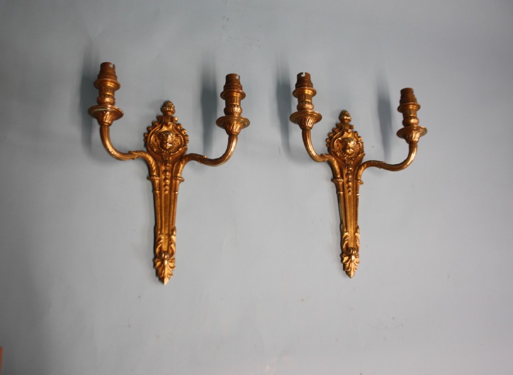 A pair of early 20th-century cast brass twin-arm wall lights featuring a cherub face motif and detailed foliate scrollwork.
