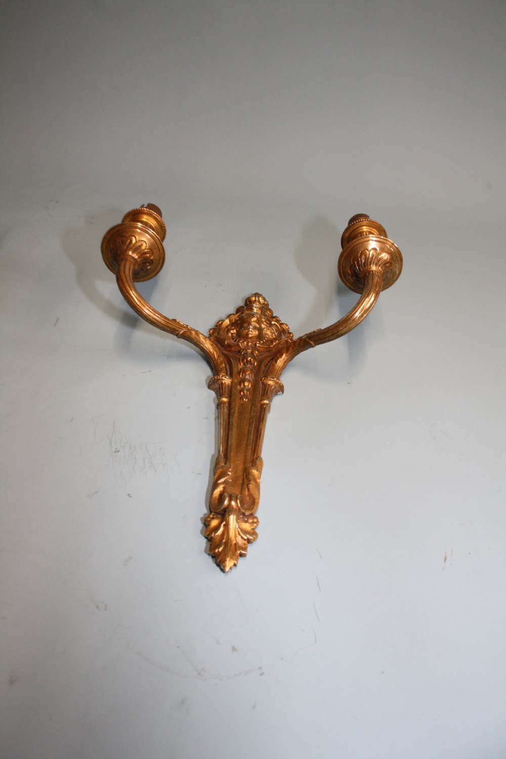 A pair of early 20th-century cast brass twin-arm wall lights featuring a cherub face motif and detailed foliate scrollwork. View IV