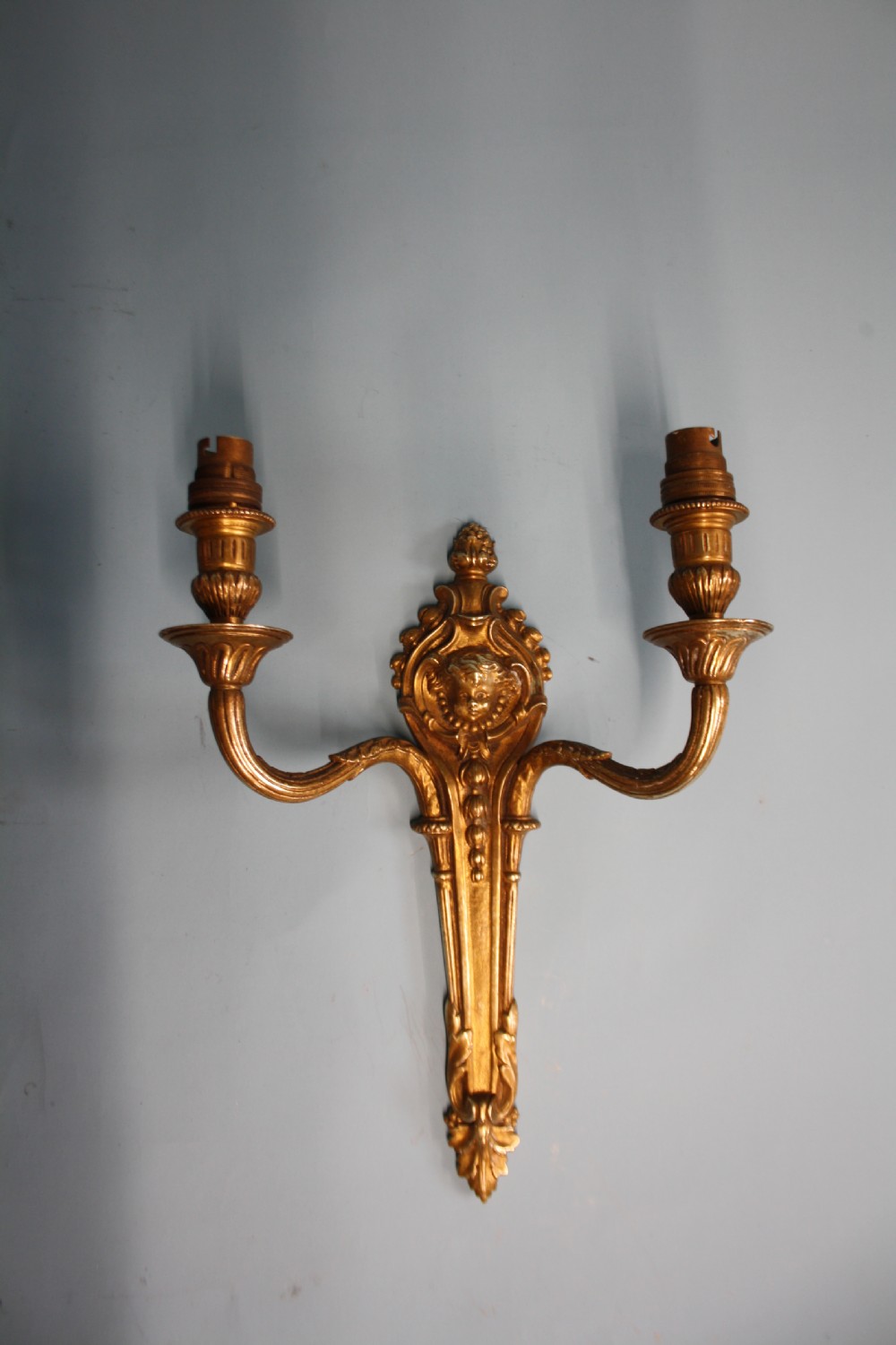 A pair of early 20th-century cast brass twin-arm wall lights featuring a cherub face motif and detailed foliate scrollwork. View X
