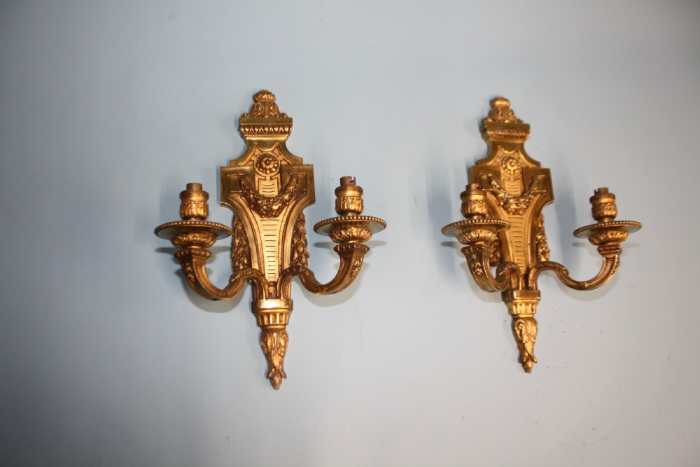A pair of early 20th-century cast brass twin-arm wall sconces featuring Neoclassical garland and wreath embellishments.