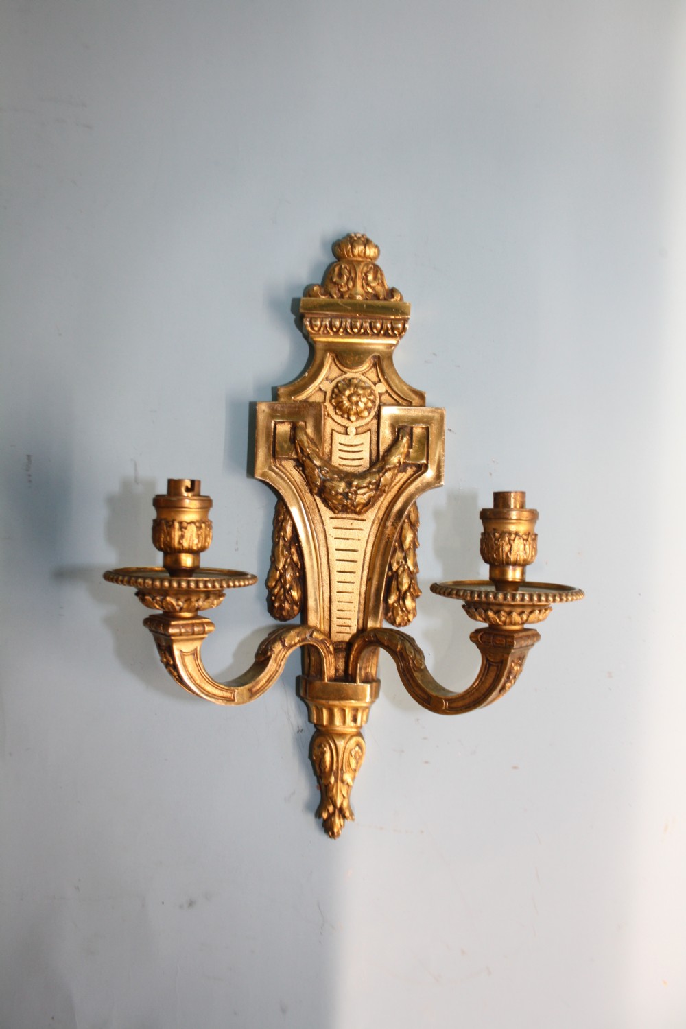 A pair of early 20th-century cast brass twin-arm wall sconces featuring Neoclassical garland and wreath embellishments. View X