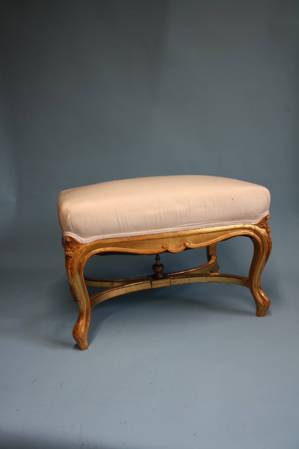 A mid-19th-century upholstered giltwood stool featuring carved cabriole legs, a serpentine seat rail, an X-shaped stretcher with a central finial, and a cream fabric top.