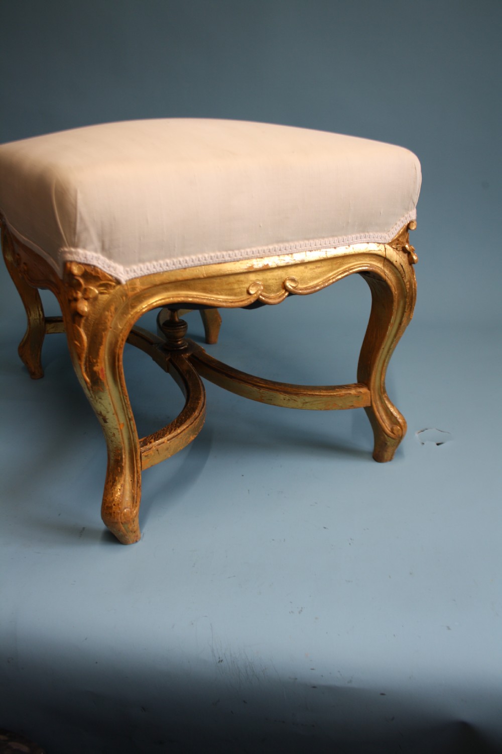 A mid-19th-century upholstered giltwood stool featuring carved cabriole legs, a serpentine seat rail, an X-shaped stretcher with a central finial, and a cream fabric top. View II