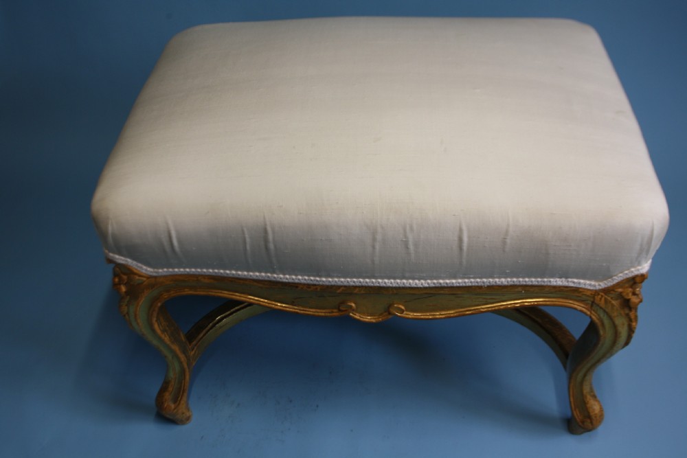A mid-19th-century upholstered giltwood stool featuring carved cabriole legs, a serpentine seat rail, an X-shaped stretcher with a central finial, and a cream fabric top. View V