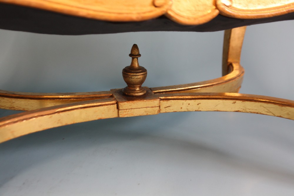 A mid-19th-century upholstered giltwood stool featuring carved cabriole legs, a serpentine seat rail, an X-shaped stretcher with a central finial, and a cream fabric top. View VI