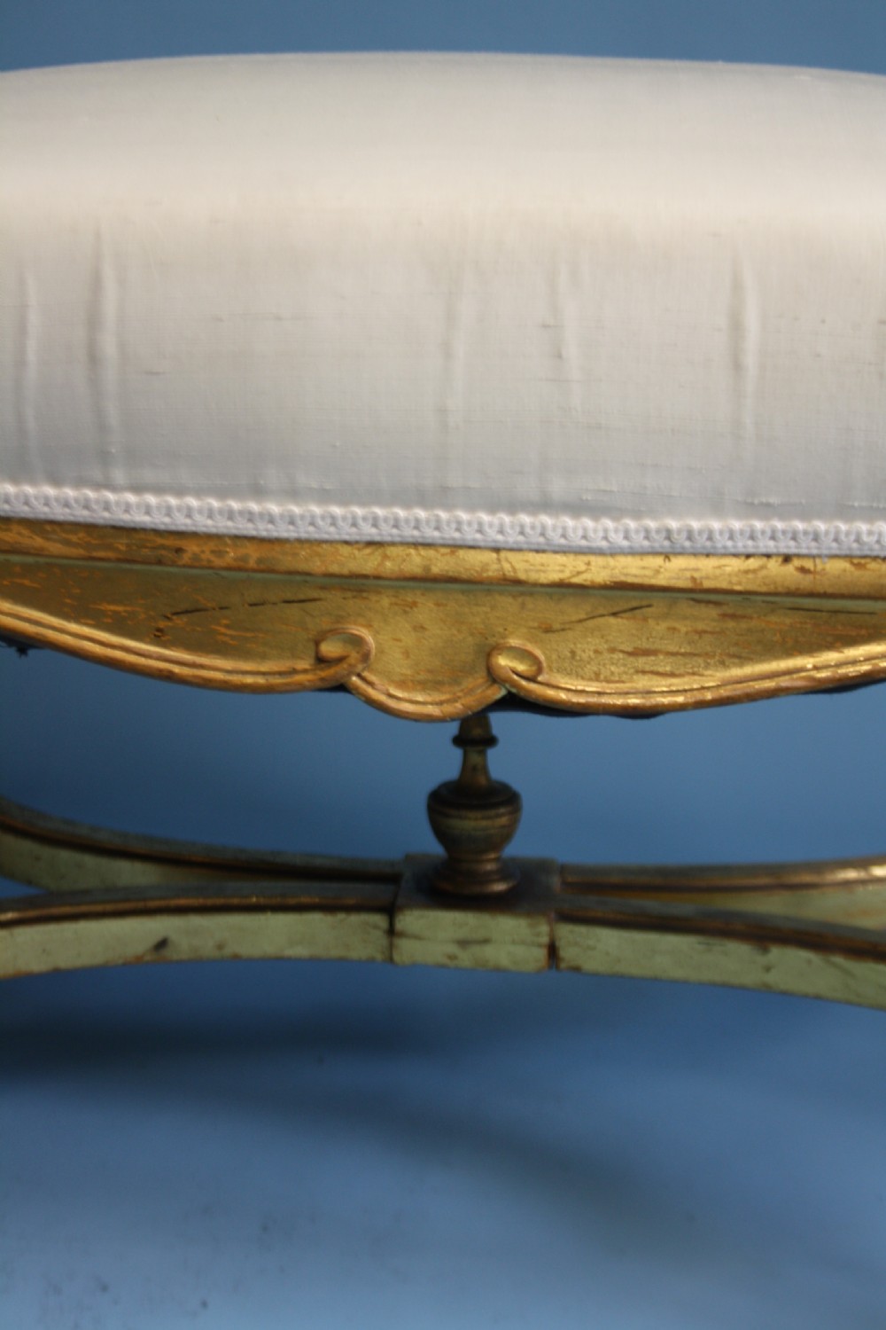 A mid-19th-century upholstered giltwood stool featuring carved cabriole legs, a serpentine seat rail, an X-shaped stretcher with a central finial, and a cream fabric top. View VII