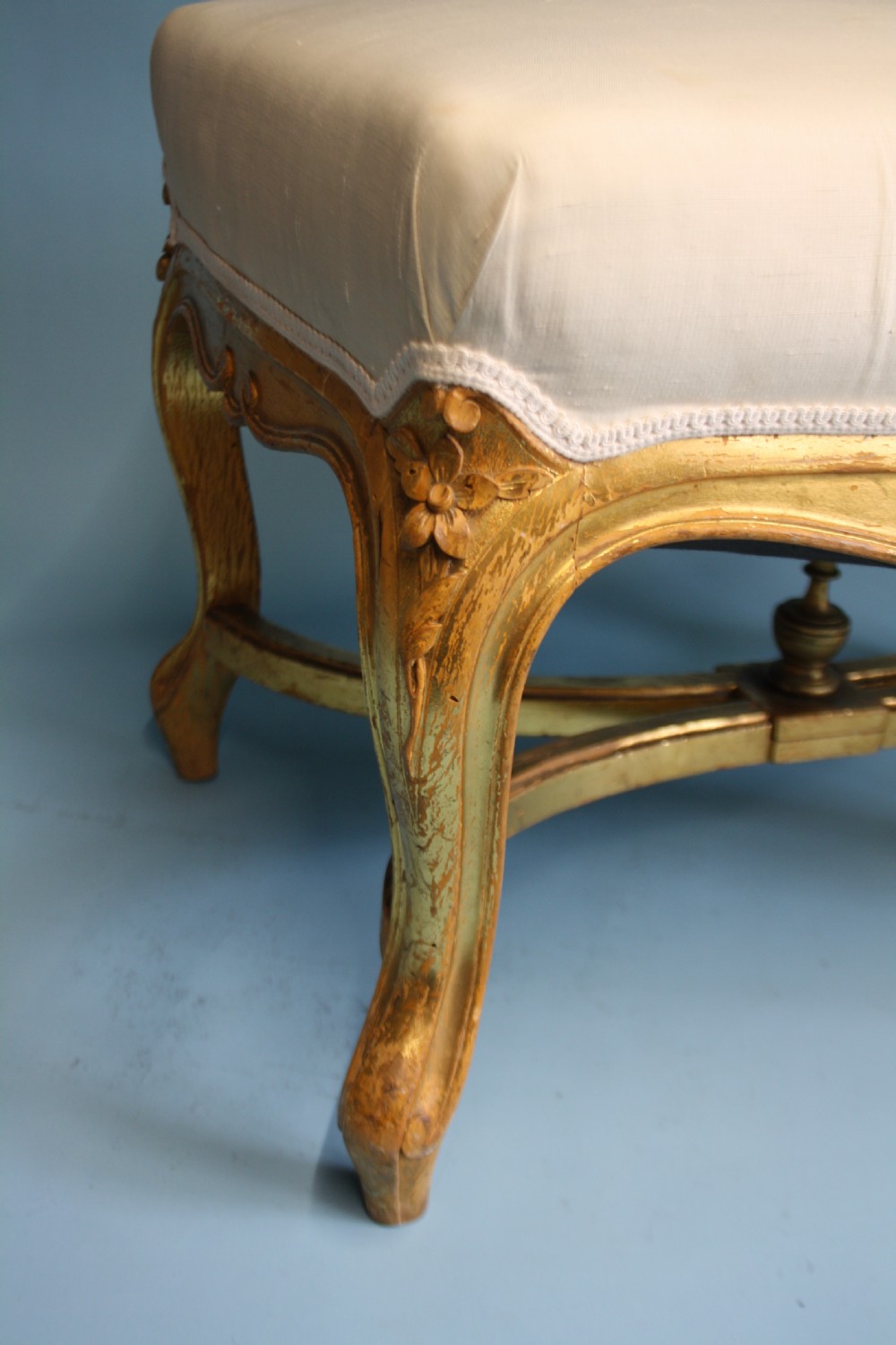 A mid-19th-century upholstered giltwood stool featuring carved cabriole legs, a serpentine seat rail, an X-shaped stretcher with a central finial, and a cream fabric top. View VIII