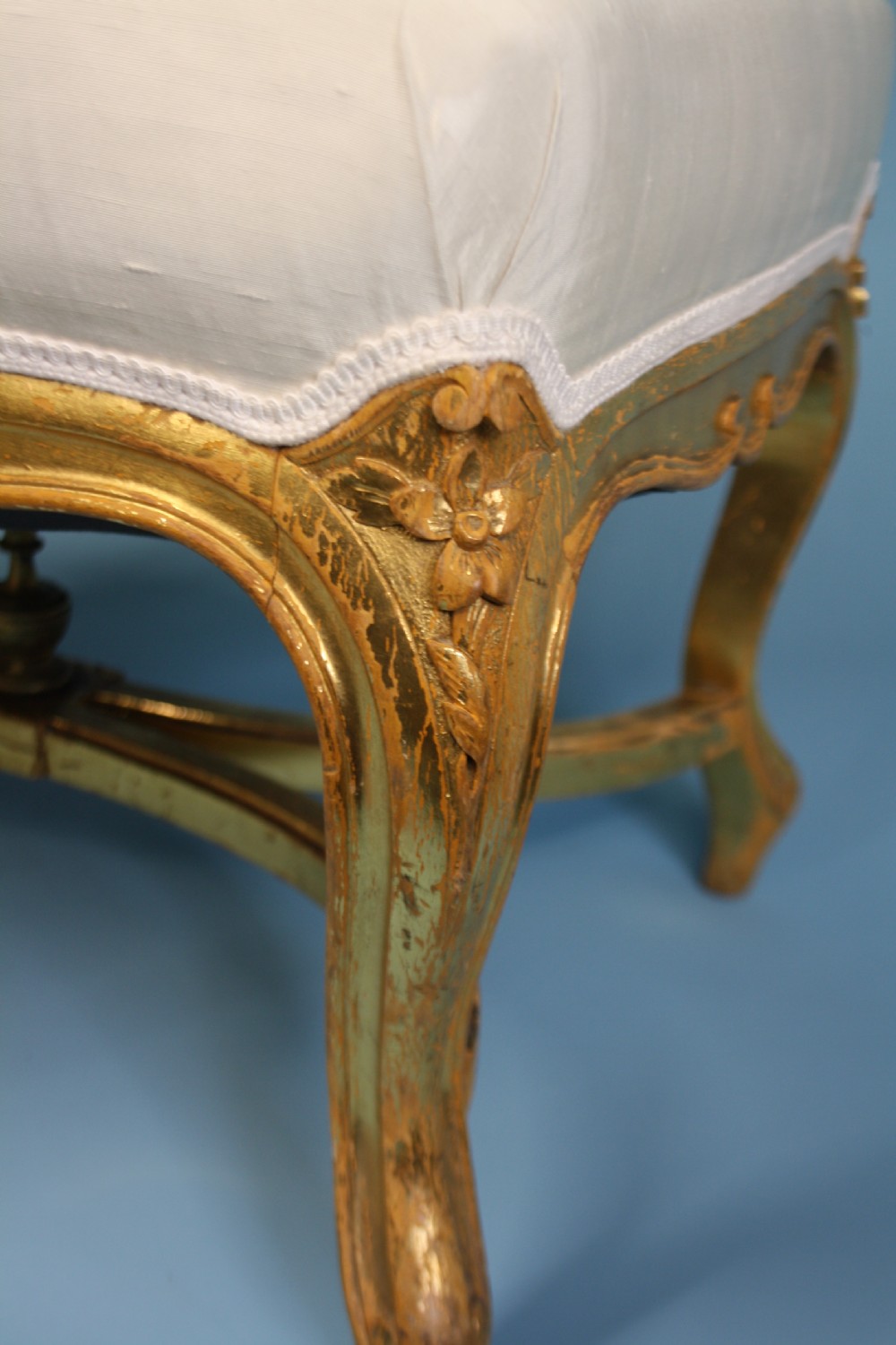 A mid-19th-century upholstered giltwood stool featuring carved cabriole legs, a serpentine seat rail, an X-shaped stretcher with a central finial, and a cream fabric top. View X