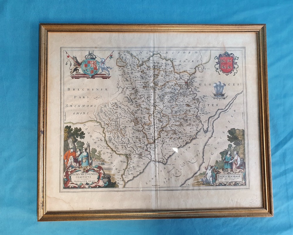 A framed 17th-century hand-coloured map of Monmouthshire featuring coats of arms, figures, a decorative cartouche, and a gilded wood frame.