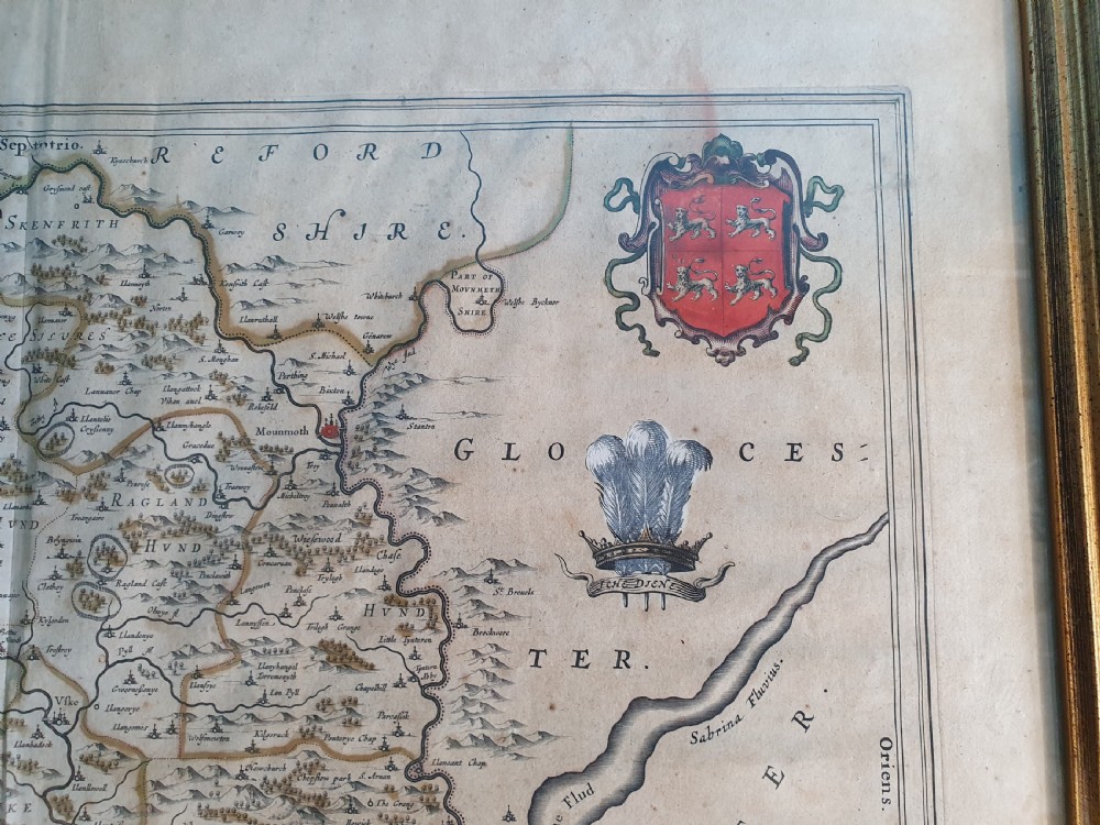 A framed 17th-century hand-coloured map of Monmouthshire featuring coats of arms, figures, a decorative cartouche, and a gilded wood frame. View V