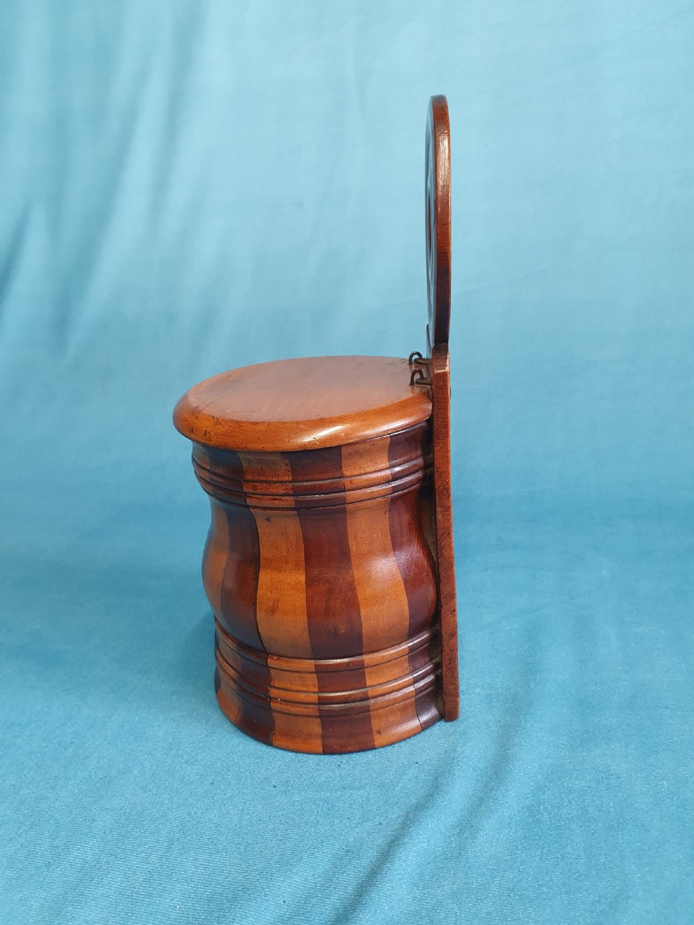 A mid-19th-century turned wooden wall-hanging salt box with staved multi-tone timber construction, a circular hinged lid, and a pierced round backboard. View II