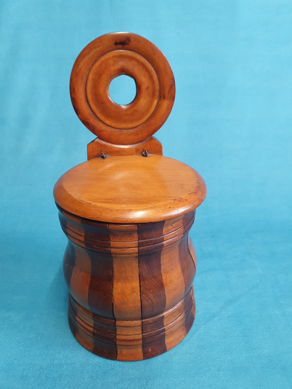 A mid-19th-century turned wooden wall-hanging salt box with staved multi-tone timber construction, a circular hinged lid, and a pierced round backboard.