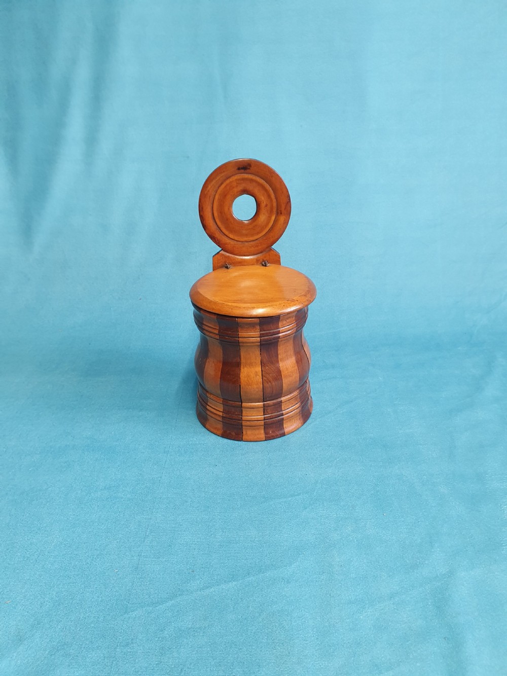 A mid-19th-century turned wooden wall-hanging salt box with staved multi-tone timber construction, a circular hinged lid, and a pierced round backboard. View VII