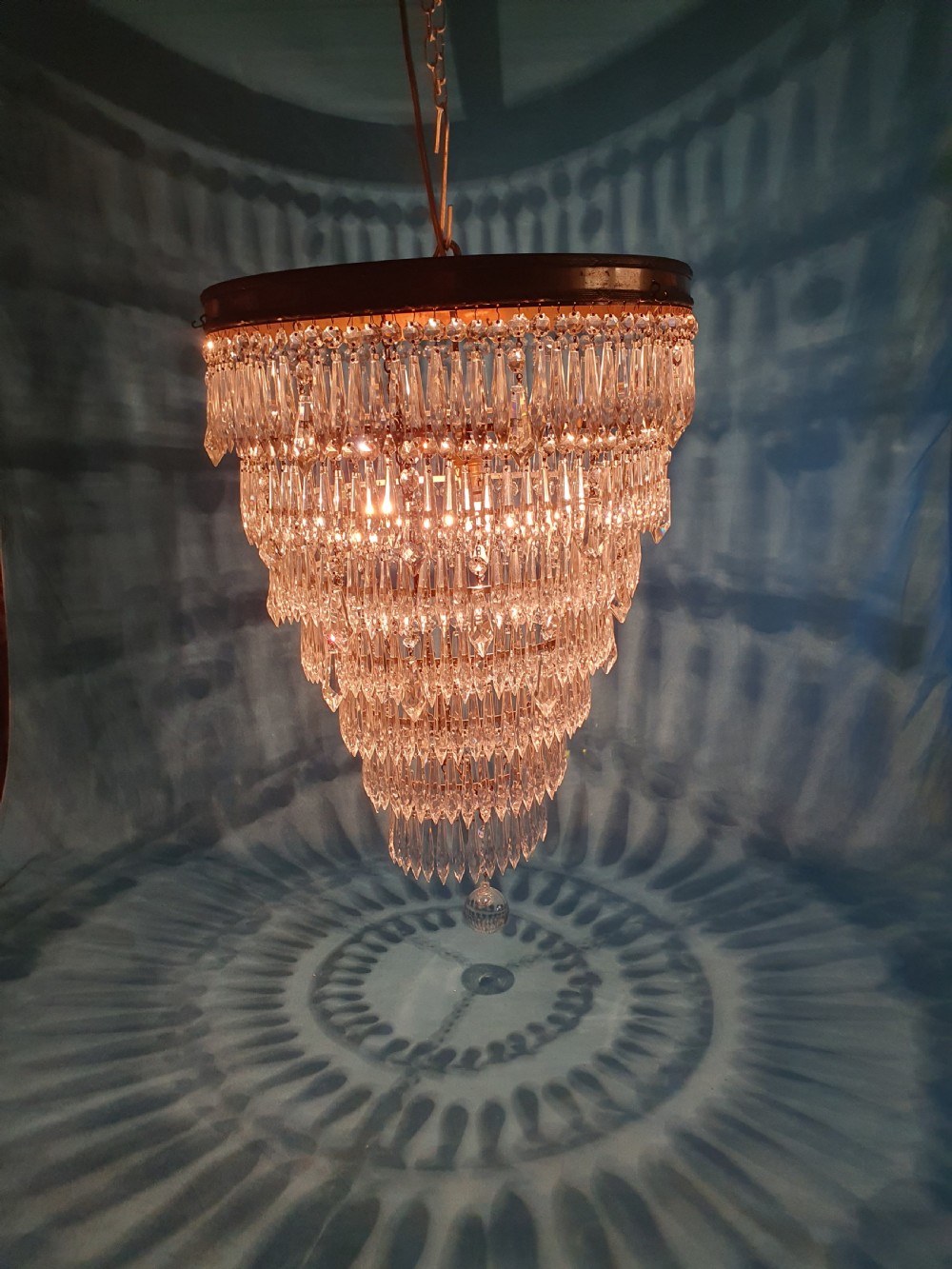A seven-tiered waterfall chandelier featuring concentric brass rings strung with elongated clear crystal spikes, ending in a single spherical glass finial. View V
