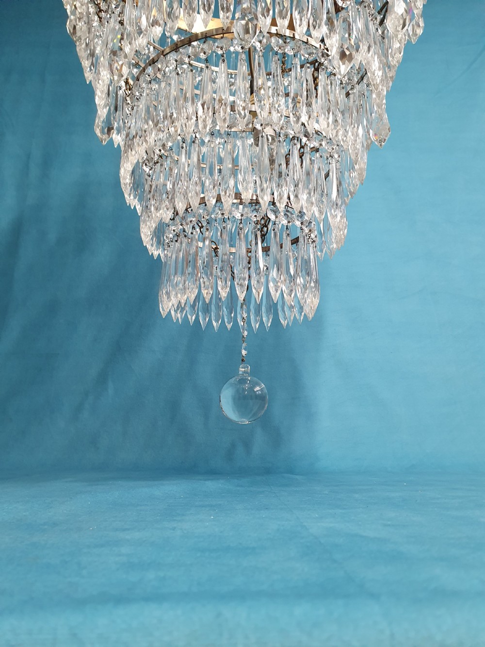 A seven-tiered waterfall chandelier featuring concentric brass rings strung with elongated clear crystal spikes, ending in a single spherical glass finial. View X