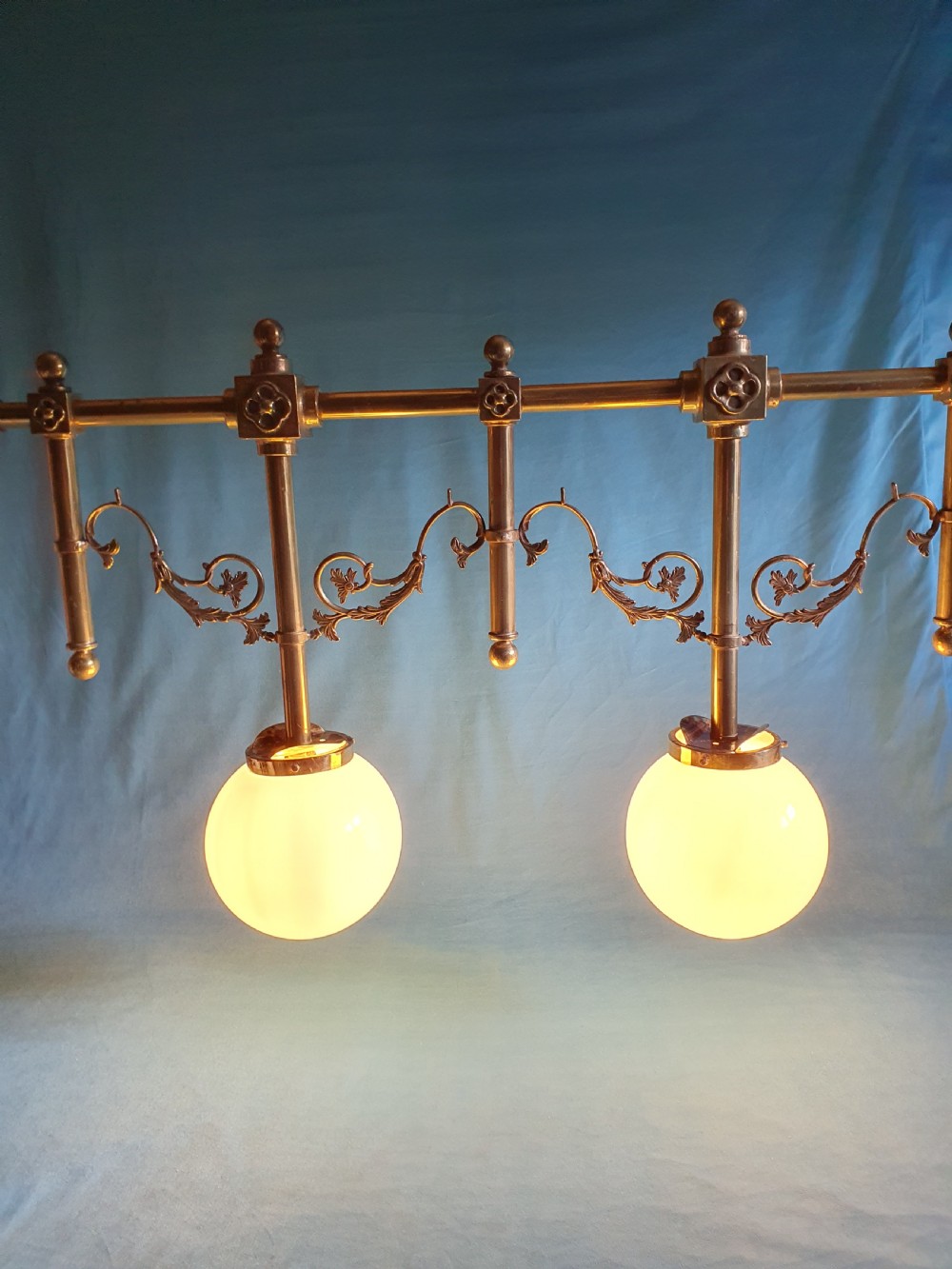 An early 20th-century Gothic Revival linear brass chandelier featuring a horizontal bar structure with ornate scrollwork brackets and two suspended white opaline glass globes. View X