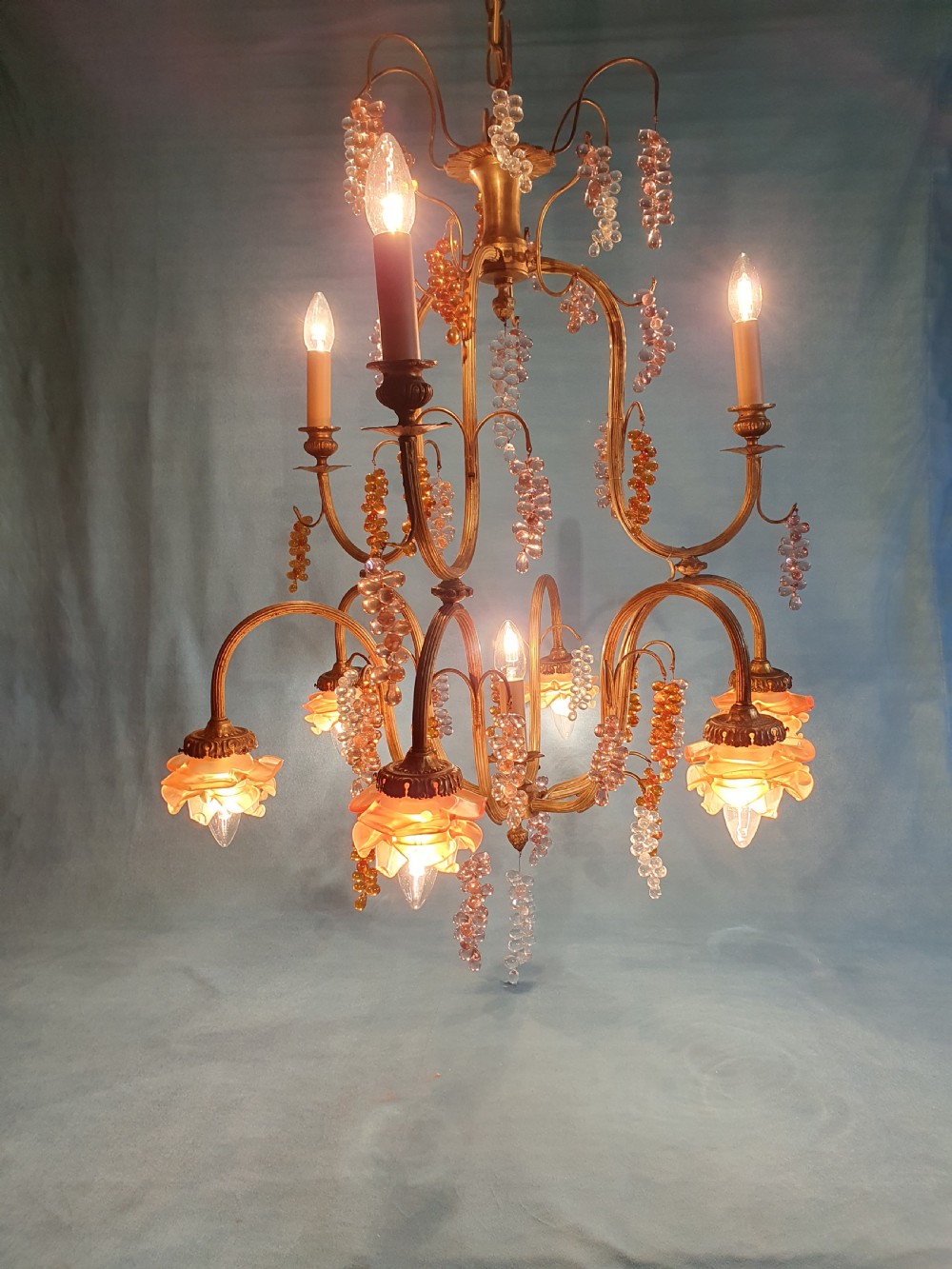A French brass chandelier adorned with multi-coloured glass grape clusters, scrollwork arms, floral glass cups, and white lampshades. View V