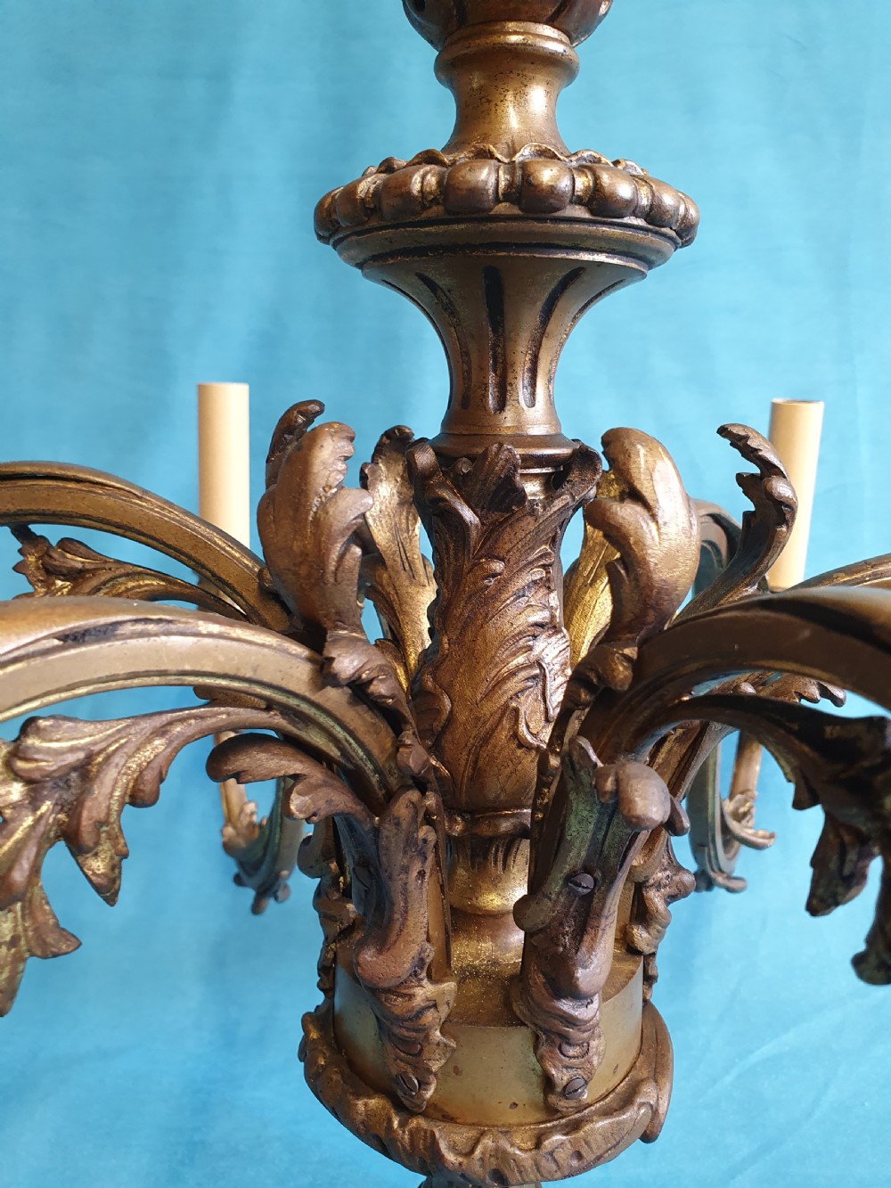 A six-arm cast brass French chandelier featuring scrollwork branches, central acanthus leaf detailing, and small white pleated fabric lampshades. View VII