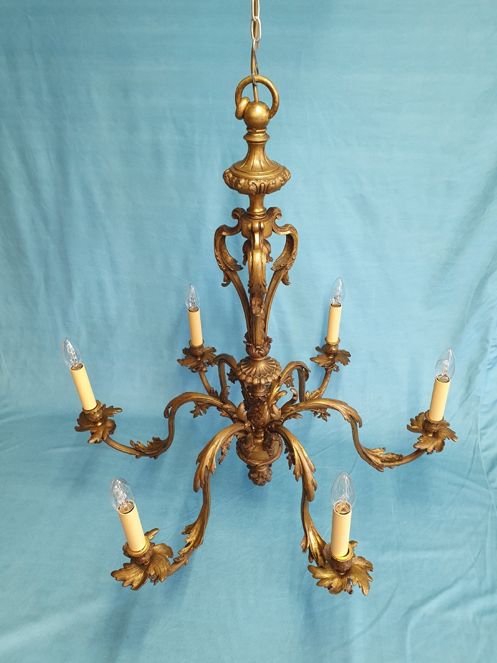 A six-arm cast brass French chandelier featuring scrollwork branches, central acanthus leaf detailing, and small white pleated fabric lampshades. View IX