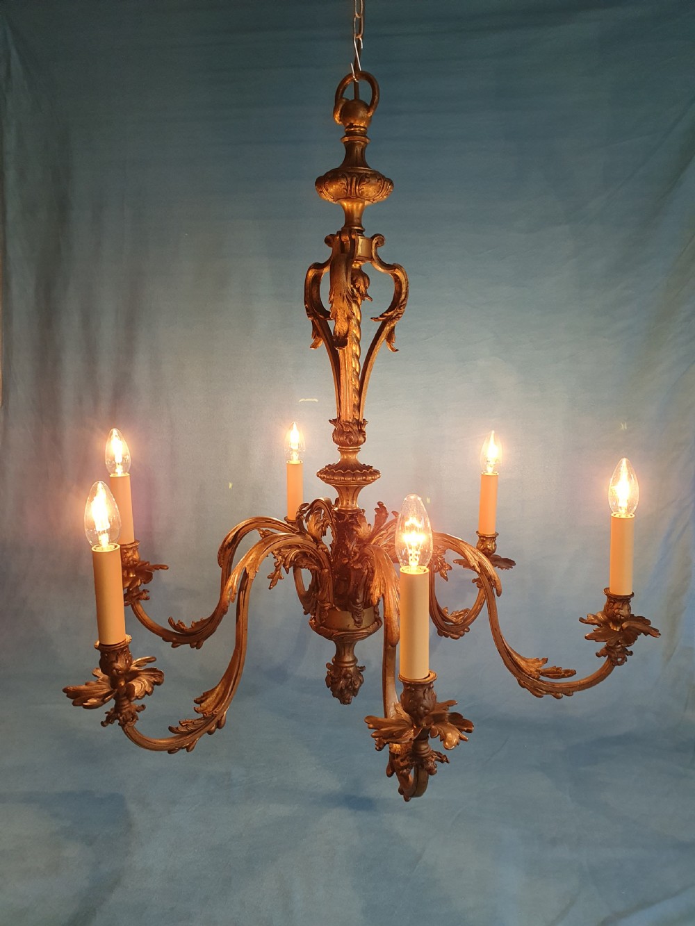A six-arm cast brass French chandelier featuring scrollwork branches, central acanthus leaf detailing, and small white pleated fabric lampshades. View X