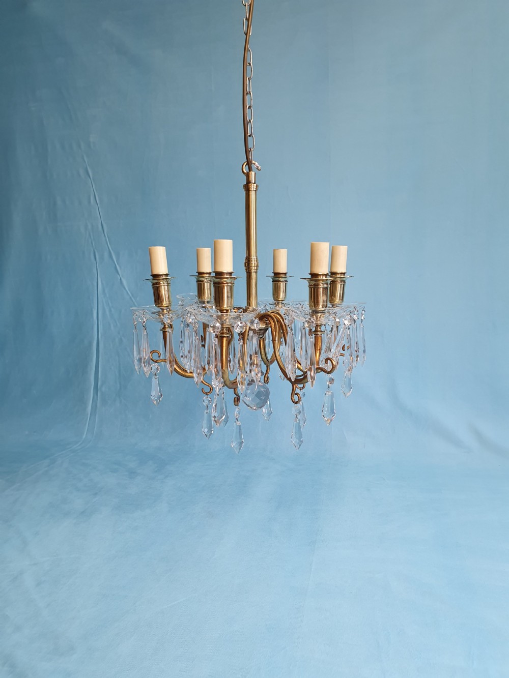 A small six-arm brass chandelier featuring glass drip pans, hanging faceted crystal drops, and a central brass stem with a suspension chain.