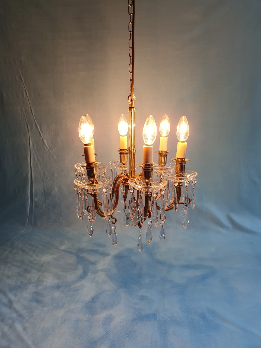 A small six-arm brass chandelier featuring glass drip pans, hanging faceted crystal drops, and a central brass stem with a suspension chain. View VII