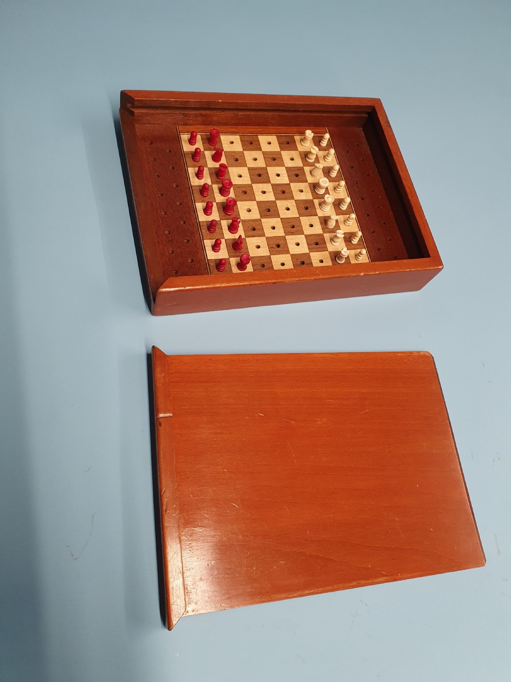An open Edwardian miniature wooden travel chess set with red and white pegged pieces set on a checkered board beside its sliding lid.