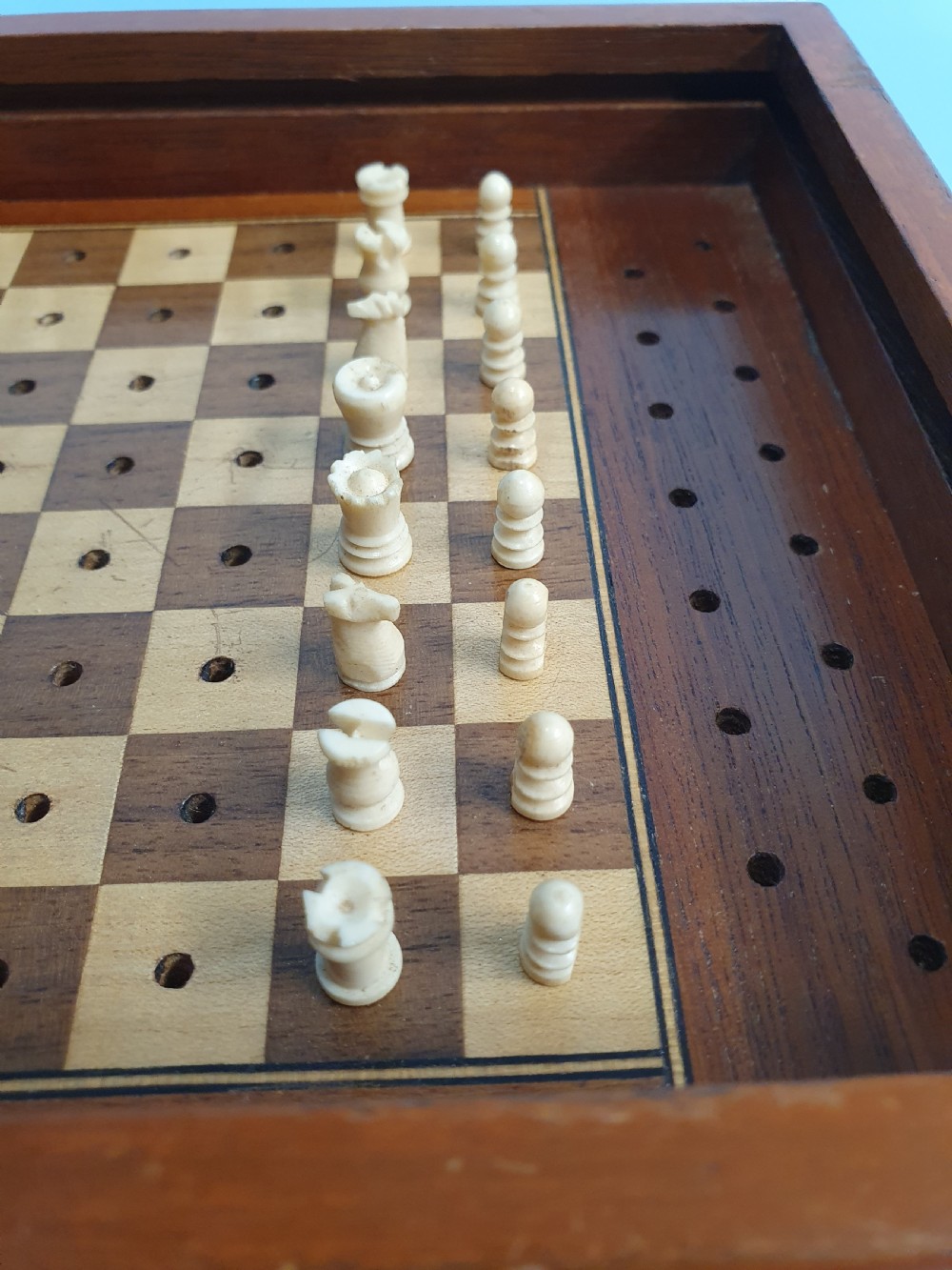 An open Edwardian miniature wooden travel chess set with red and white pegged pieces set on a checkered board beside its sliding lid. View II