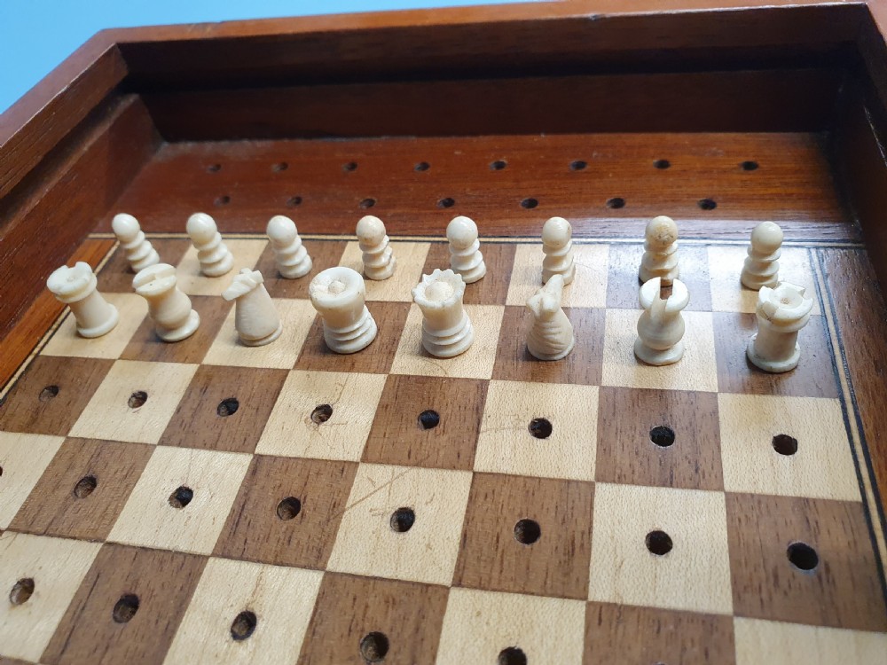 An open Edwardian miniature wooden travel chess set with red and white pegged pieces set on a checkered board beside its sliding lid. View IV