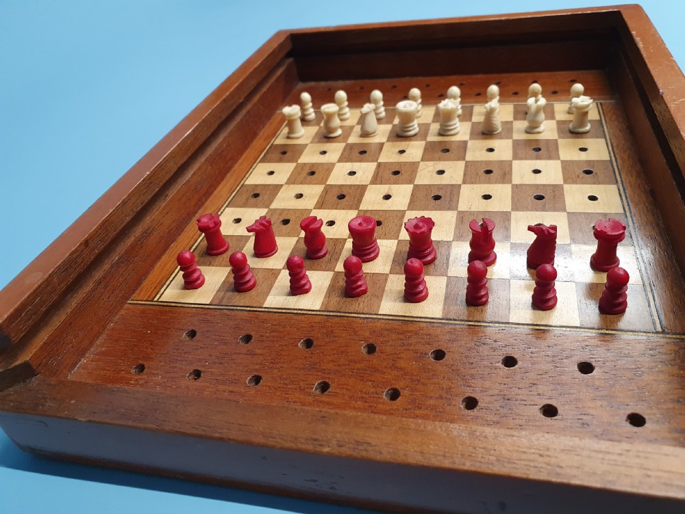 An open Edwardian miniature wooden travel chess set with red and white pegged pieces set on a checkered board beside its sliding lid. View VI