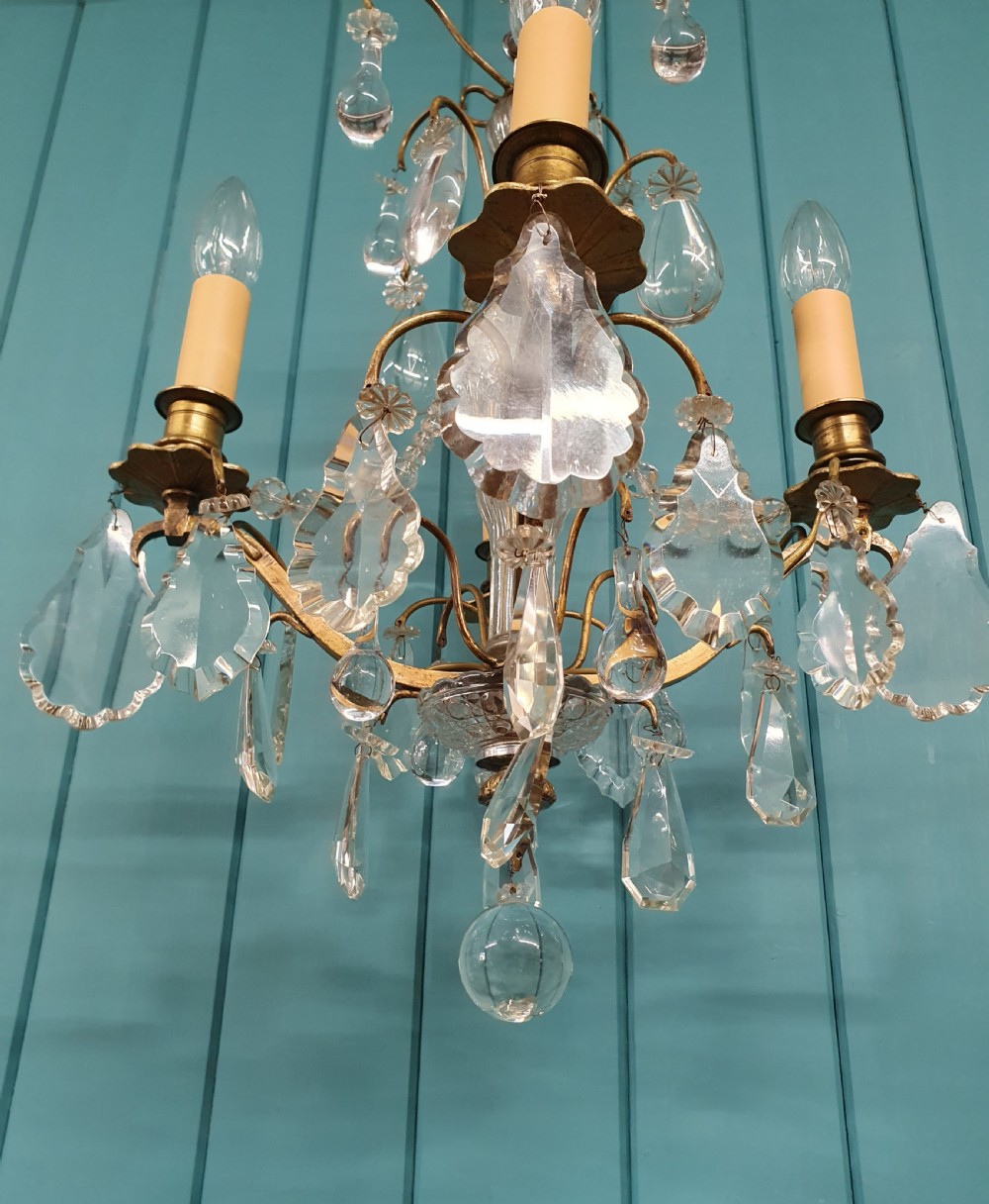 An antique French brass and crystal chandelier featuring curved scroll arms, a central glass stem, candle sleeves, and hanging teardrop glass prisms. View V